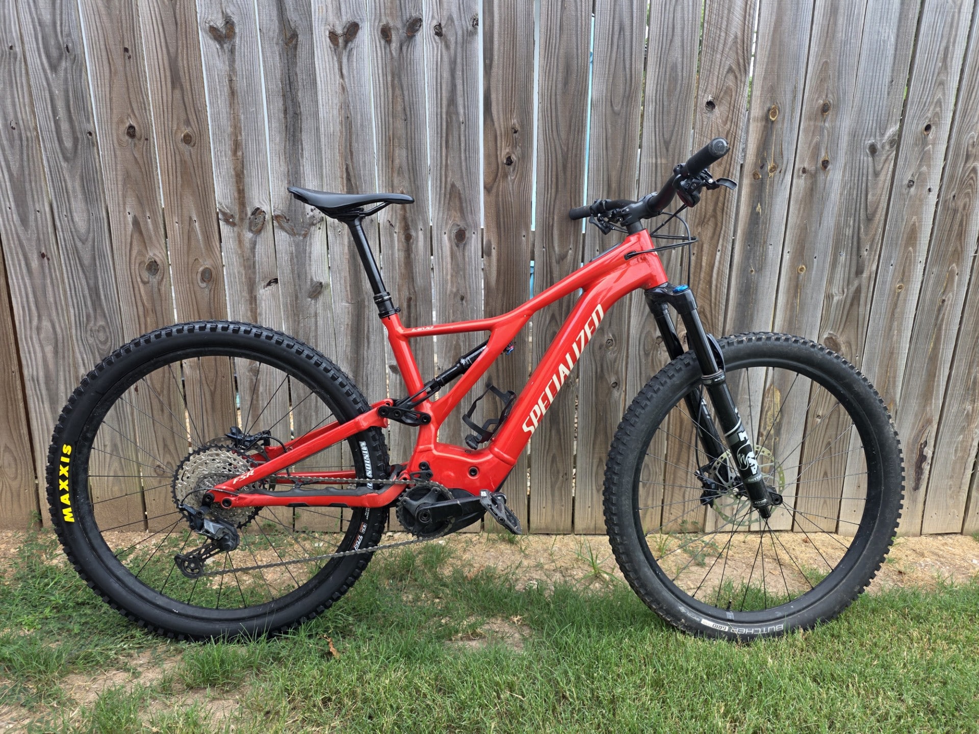 Red Specialized full-suspension mountain bike leaning against a wooden fence, side view showing knobby tires and blue front suspension cap.
