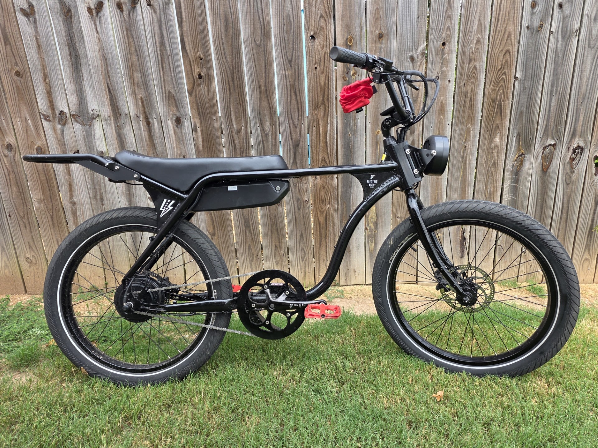 Black electric bike on a grassy lawn with a weathered wooden fence; a red cloth is draped over the handlebars.