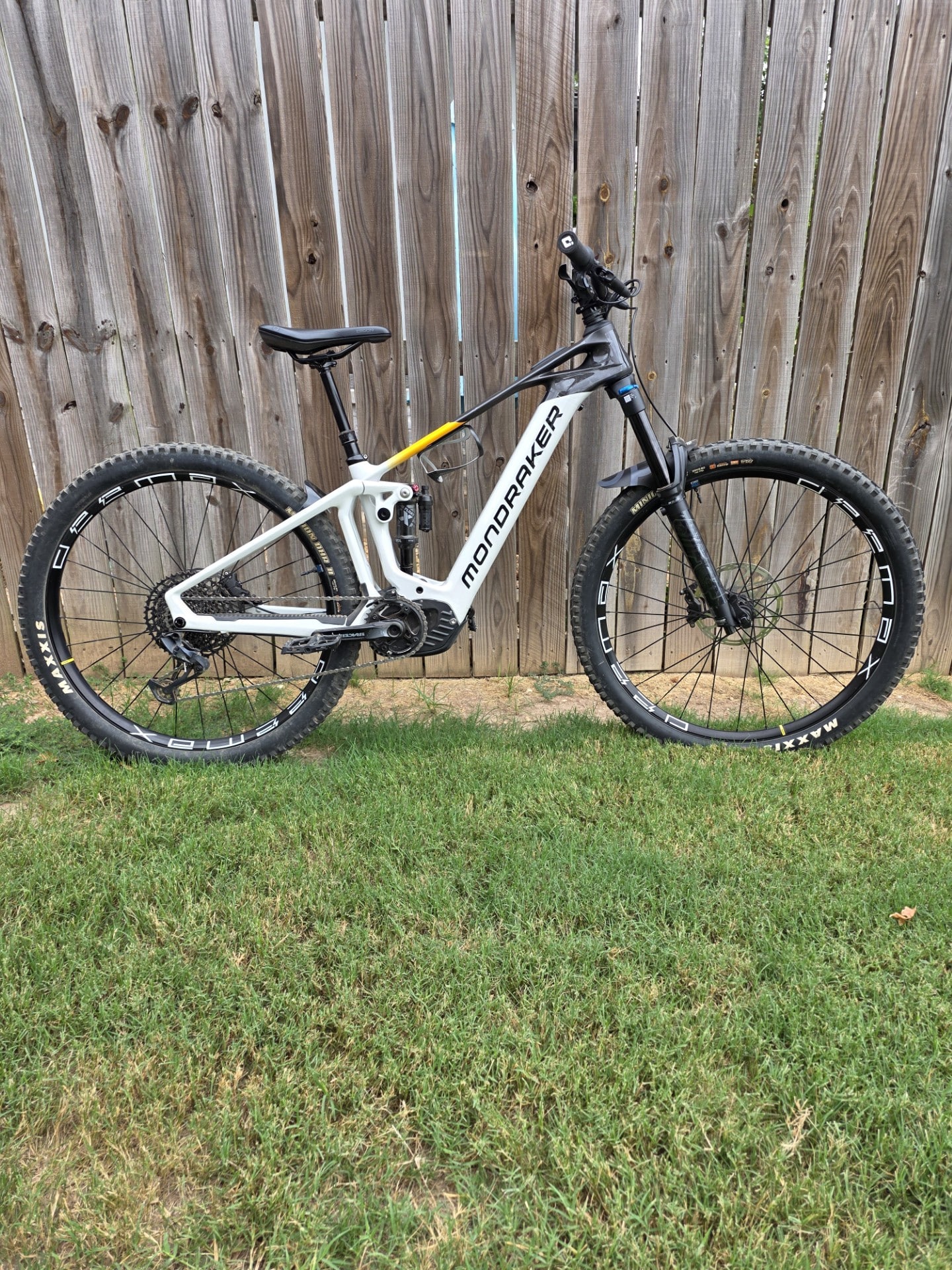 White Mondraker full-suspension mountain bike leaning against a wooden fence; black front fork, knobby Maxxis tires, and yellow accent on frame.