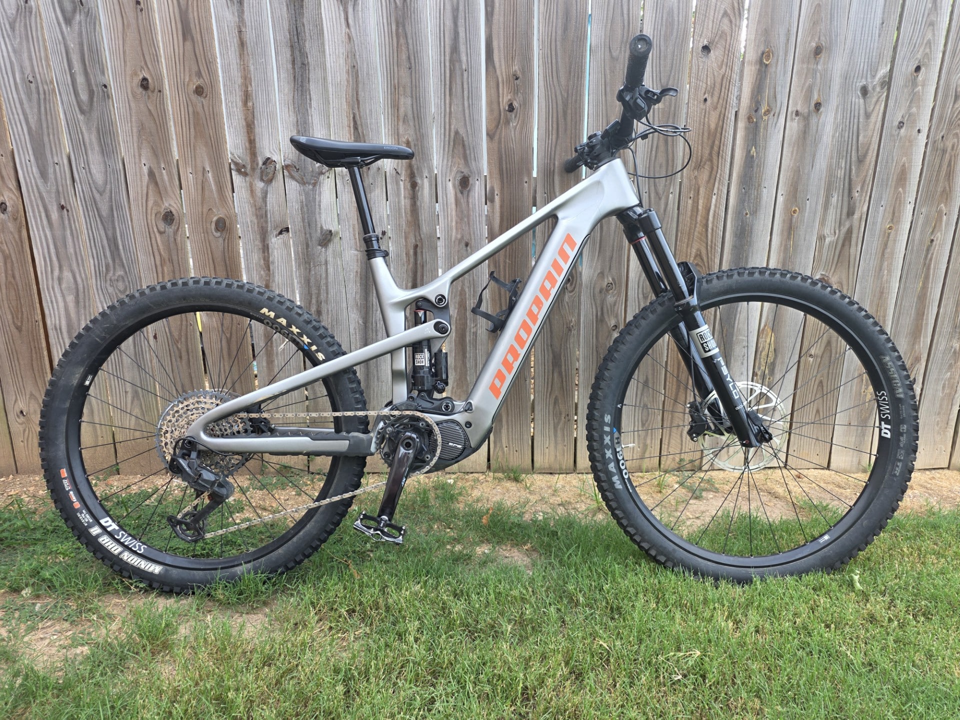 Silver full-suspension mountain bike leaning against a wooden fence, orange 'ROCKR' logo on downtube, knobby tires ready for off-road riding.